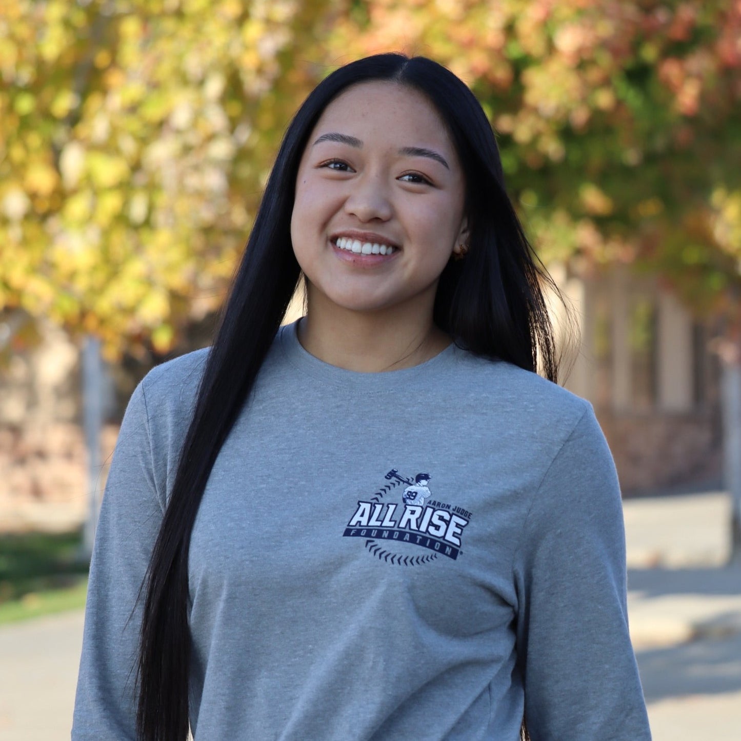 Young woman wearing a grey long sleeve tee with the All RISE logo over a baseball.